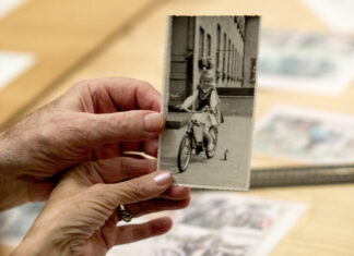 Dwie dłonie, kobieca i męska trzymają stare, czarno-białe zdjęcie. Na zdjęciu dziewczynka na w stroju ludowym na rowerku.