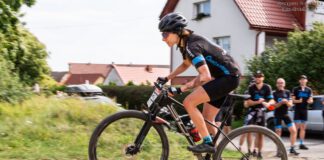 kobieta na rowerze w stroju kolarskim, w tle kibice i domy