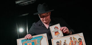 Aktor w kapeluszu trzyma w rękach karty z cyganami i wozem cygańskim