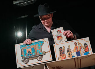 Aktor w kapeluszu trzyma w rękach karty z cyganami i wozem cygańskim