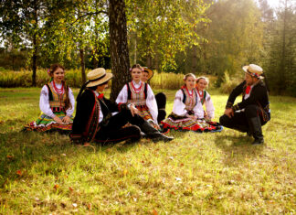 przebrani w stroje ludowe młodzi mężczyźni i kobiety siedzą na trawie